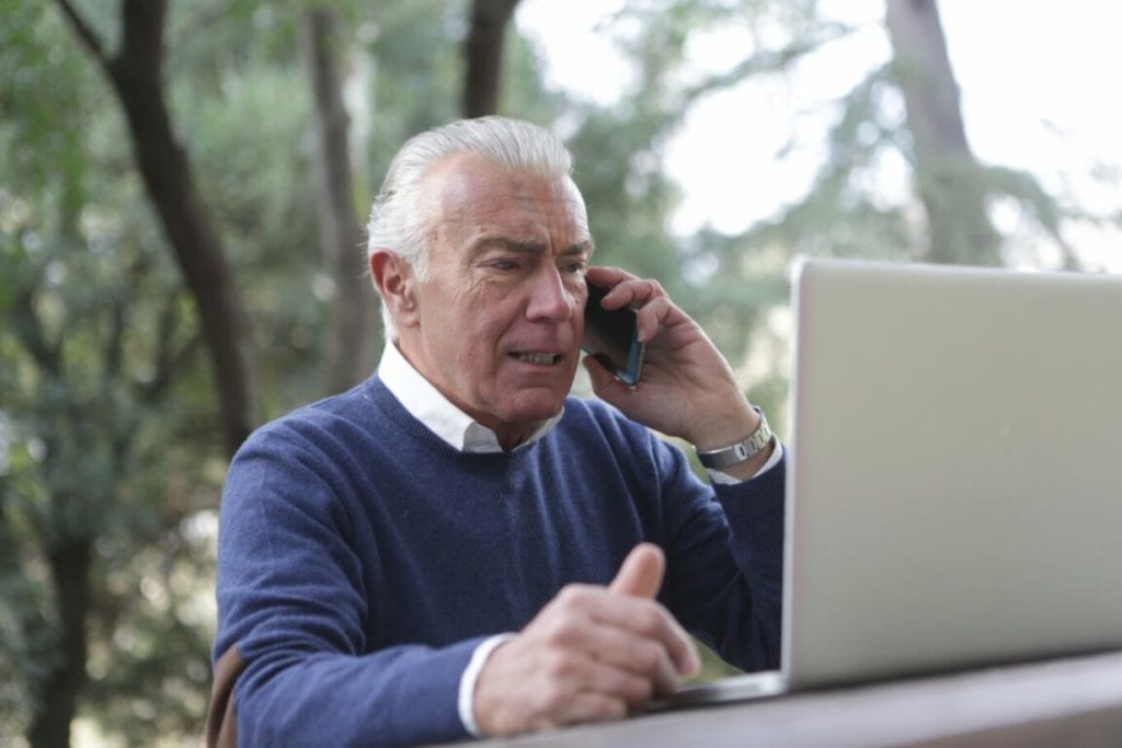an old man with with white hair and dressed in a blue cardigan talking on a phone and also using a laptop in an outdoor setting