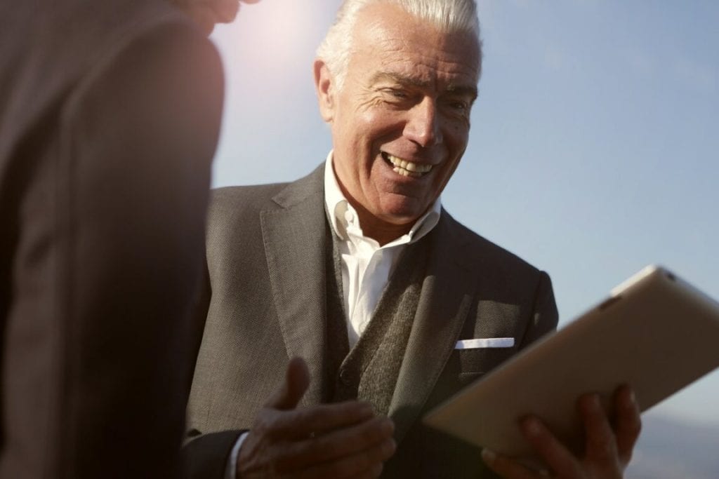 old man dressed in a dark grey suit smiling while holding a tablet