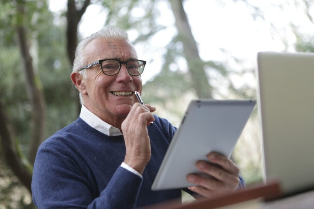 old man with grey hair smiling and holding a tablet and a pen with a laptop on a desk on an outdoor setting