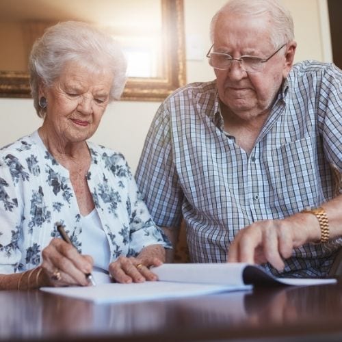 old man in checkered shirt and woman in floral jobs sitting at a wooden table. They are both looking at a document on the table with the woman signing it using a pen.