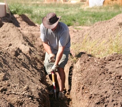 man in shorts and cow boy hat digging a trench with a shovel in the process of building a floodwall.